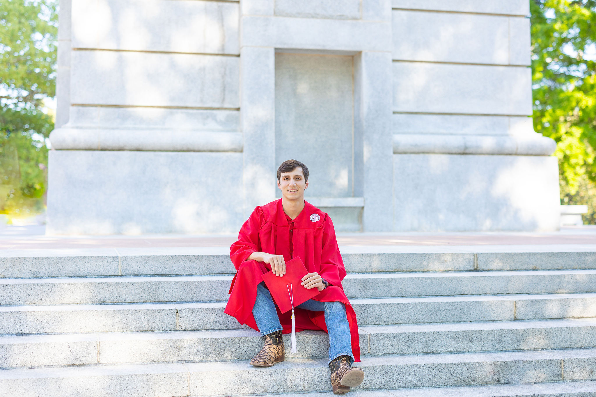 NC State seniorsitting on the belltower steps in jeans, boots, and a red gown holding his cap