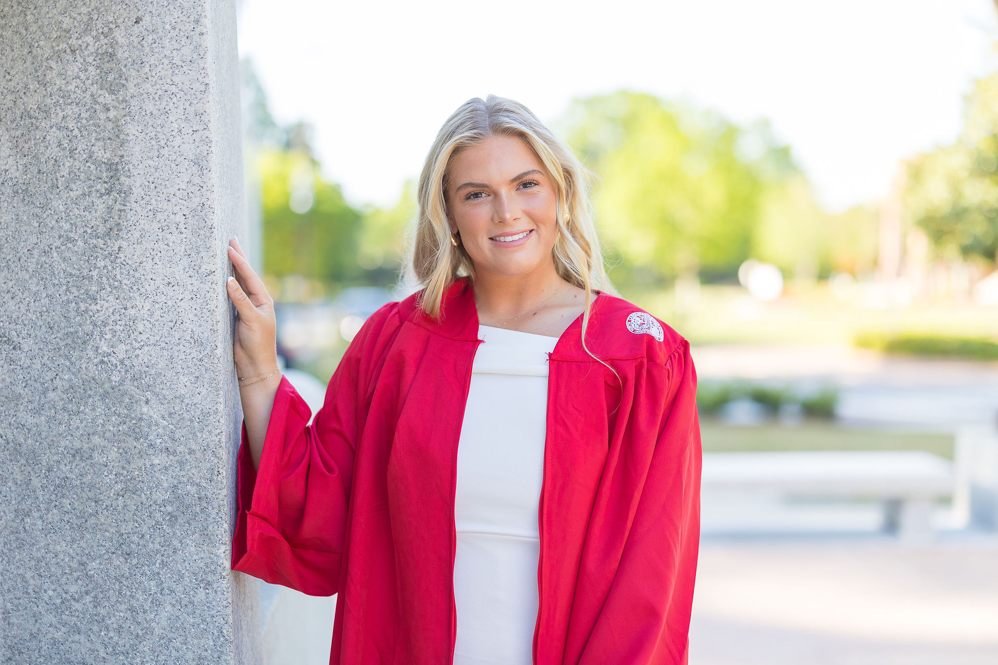 A NC State grad in a white dress and red gown standing with one hand against the belltower