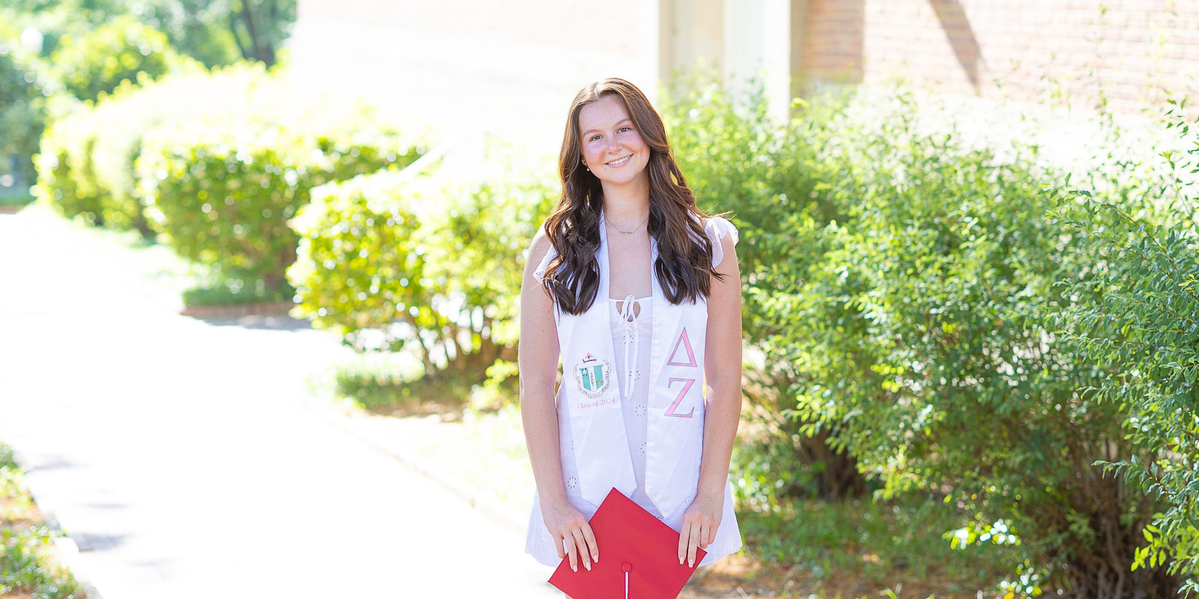 NC State Senior wearing a white dress and stole holding a red cap outside posing in front of greenery