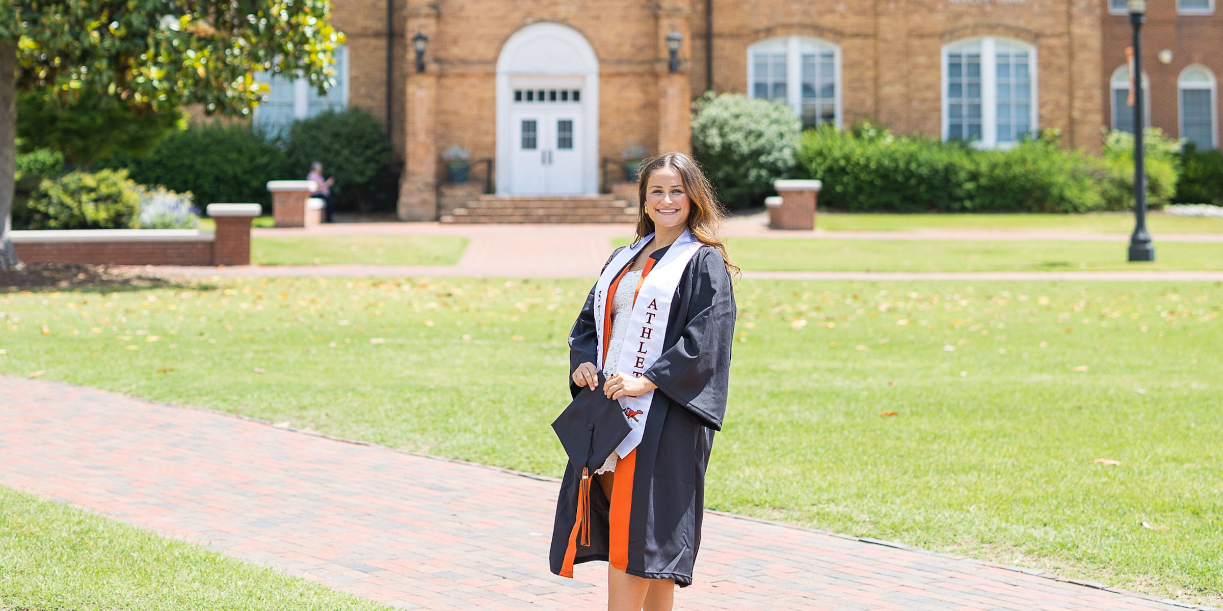 Campbell University graduate wearing a black graduation gown and a Student-Athlete stole in front of a brick building with green grass