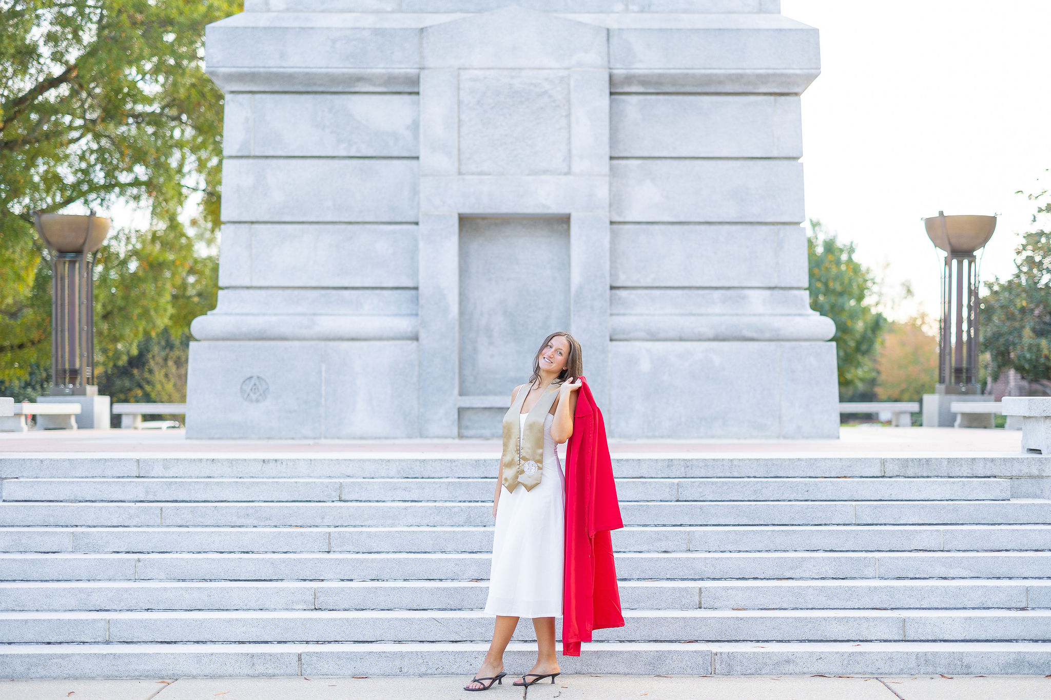 girl in white dress at the NC State Belltower holding a red graduation gown over her shoulder