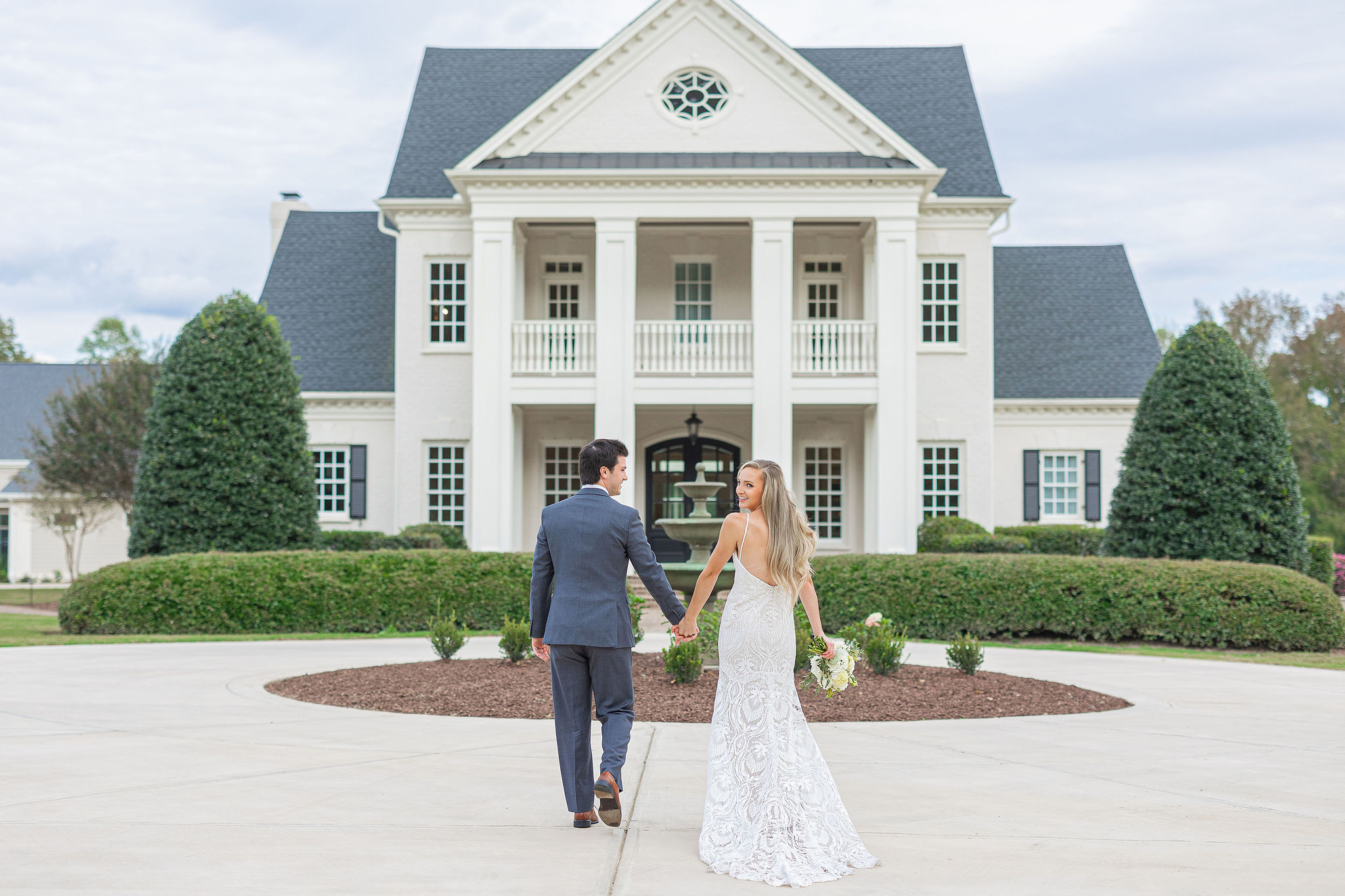 Bride and groom in front of large house holding hands while bride is looking back