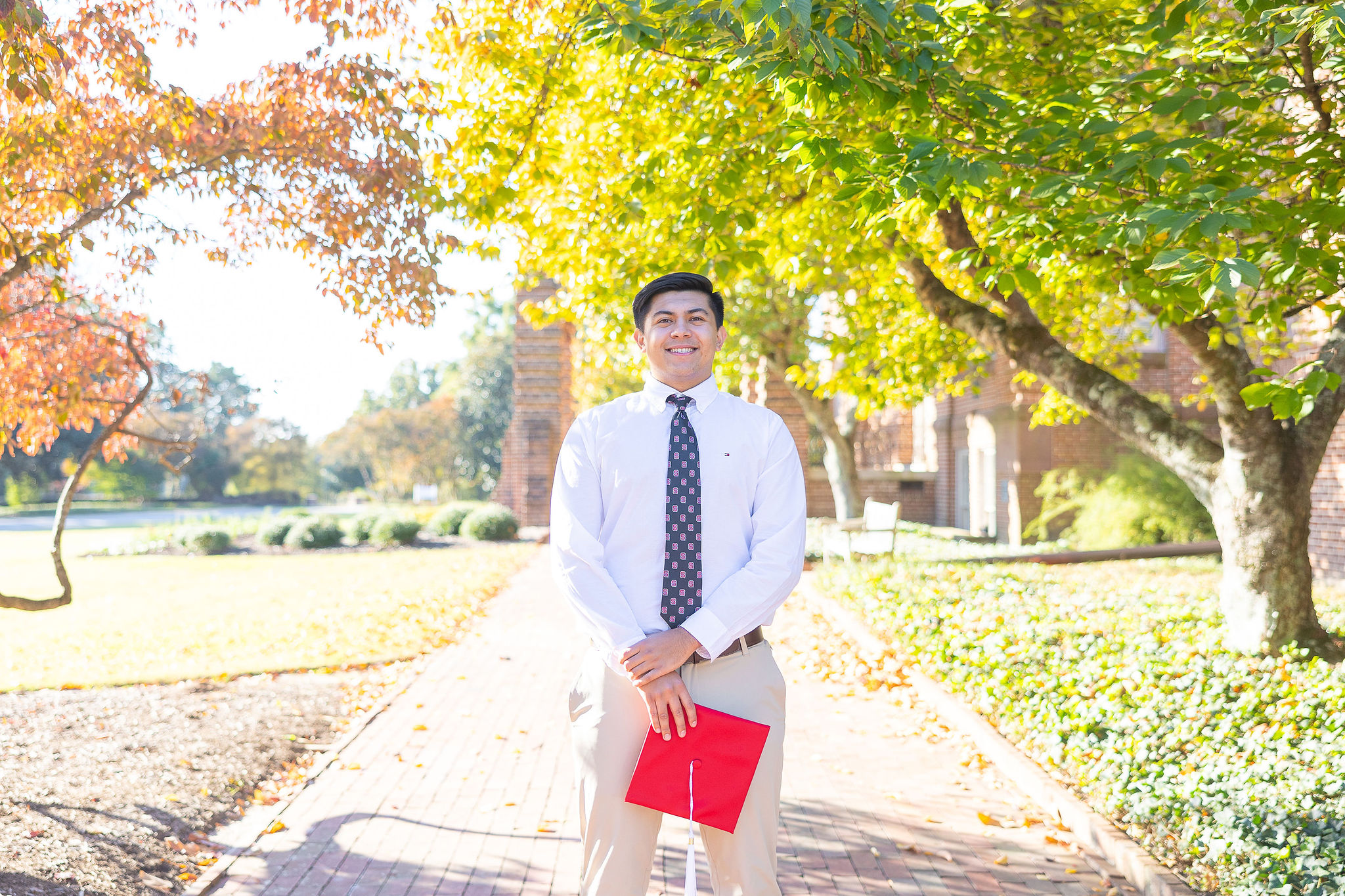 NC State male graduate standing in front of trees holding red cap with white button down and blue tie