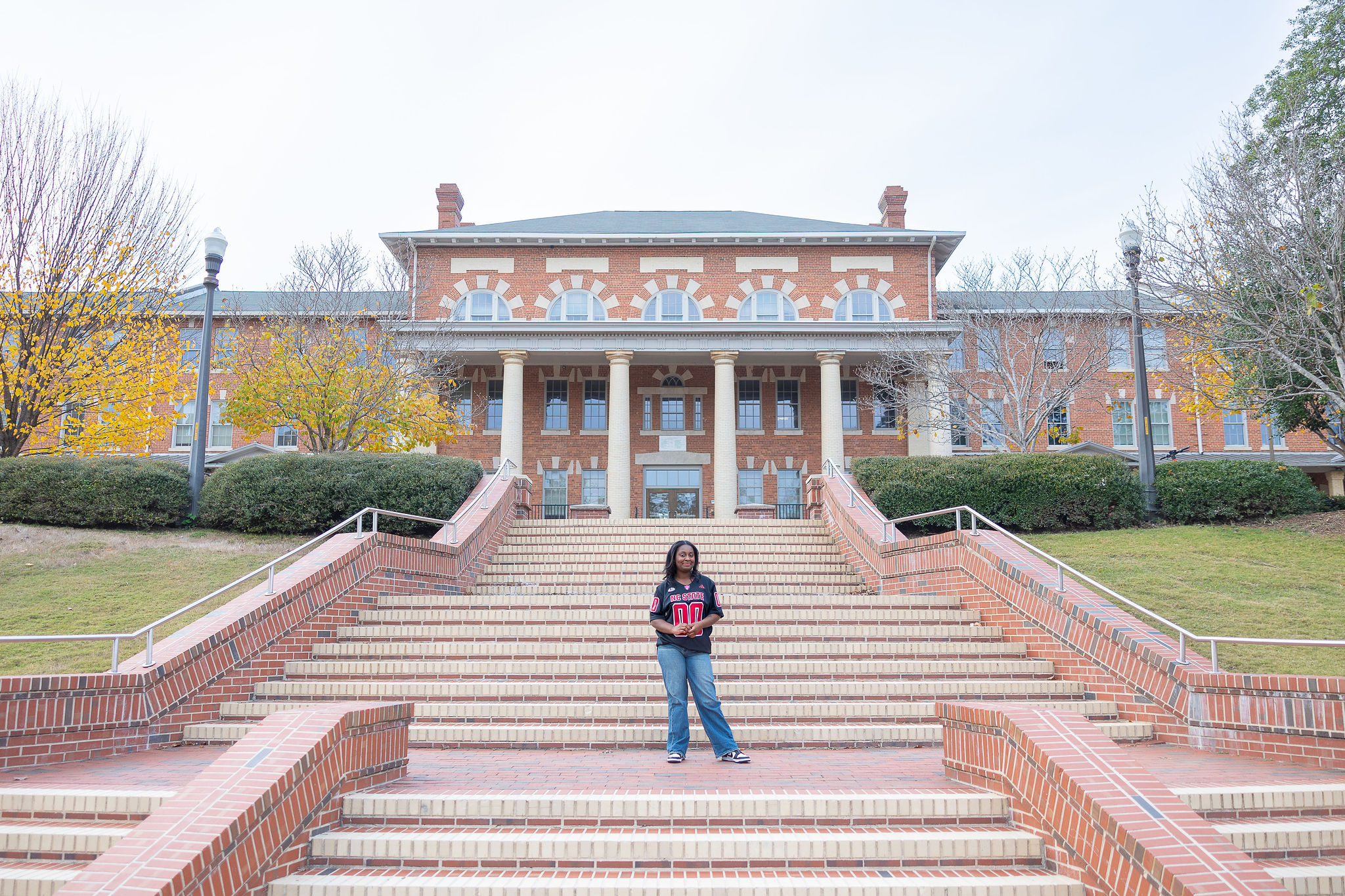 NC State grad standing on brick steps in a black football jersey and jeans