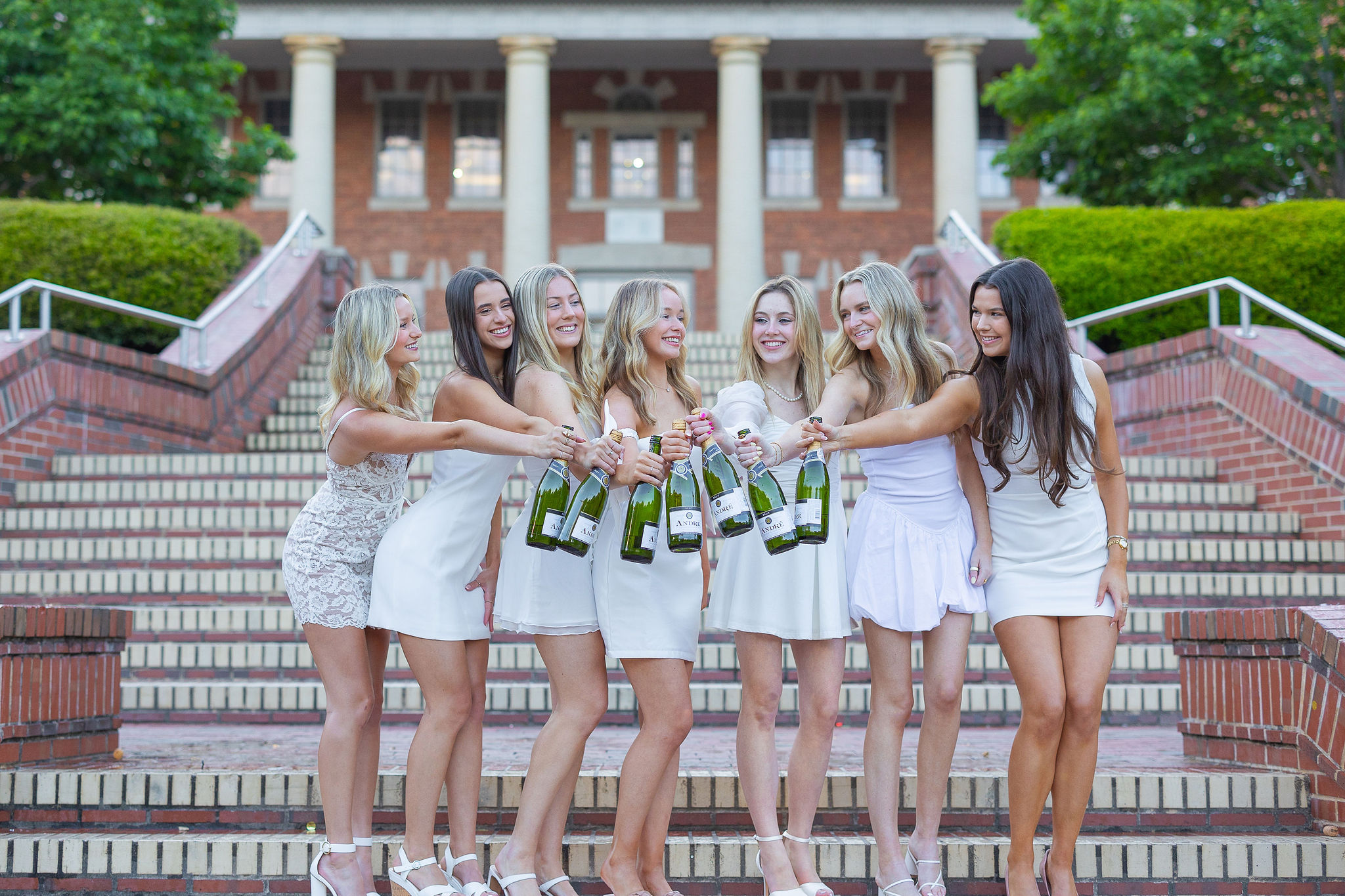 Group of girls in white dresses standing on brick steps with bottles of champagne