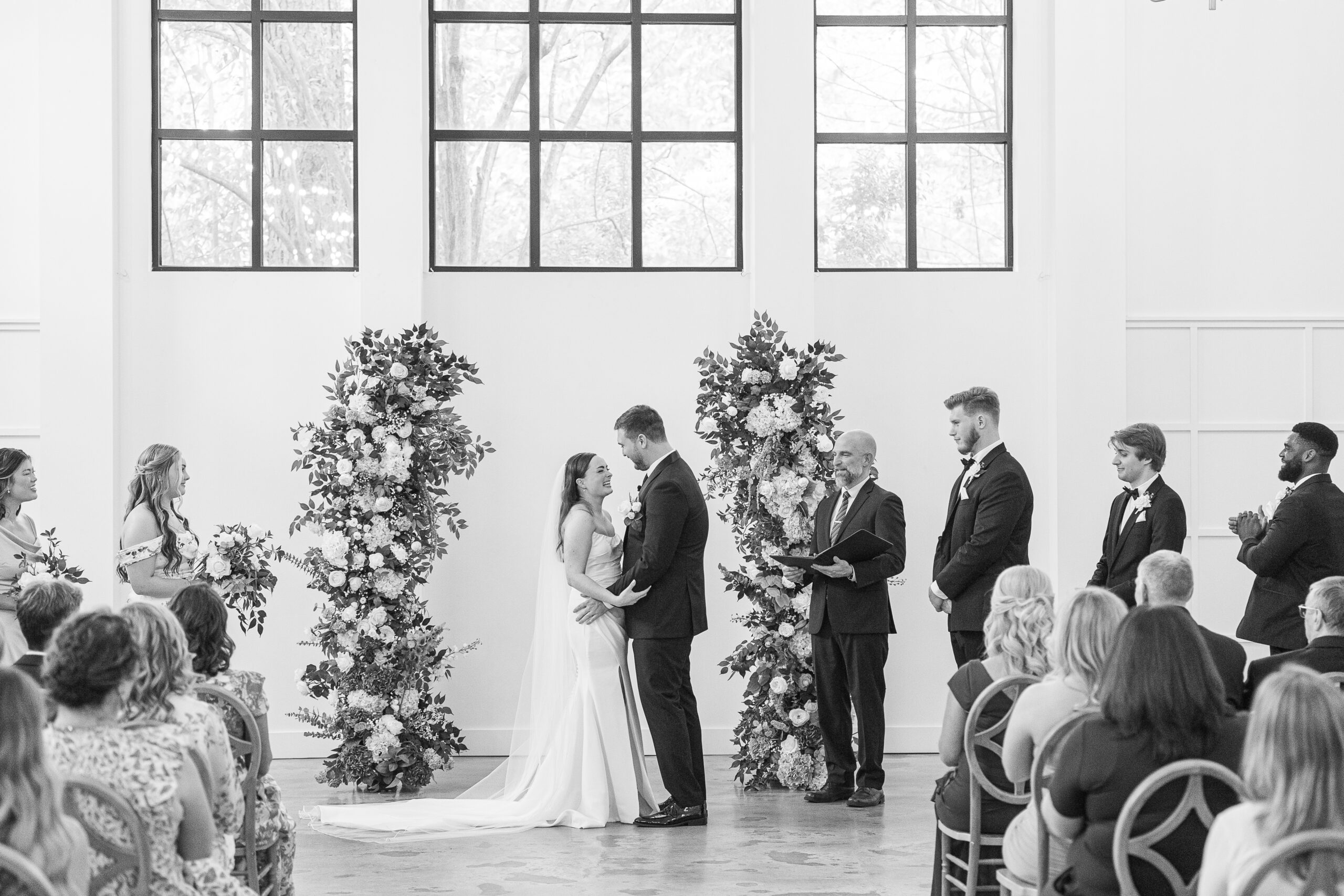 black and white photo of bride and groom at alter at indoor wedding venue with flower arch