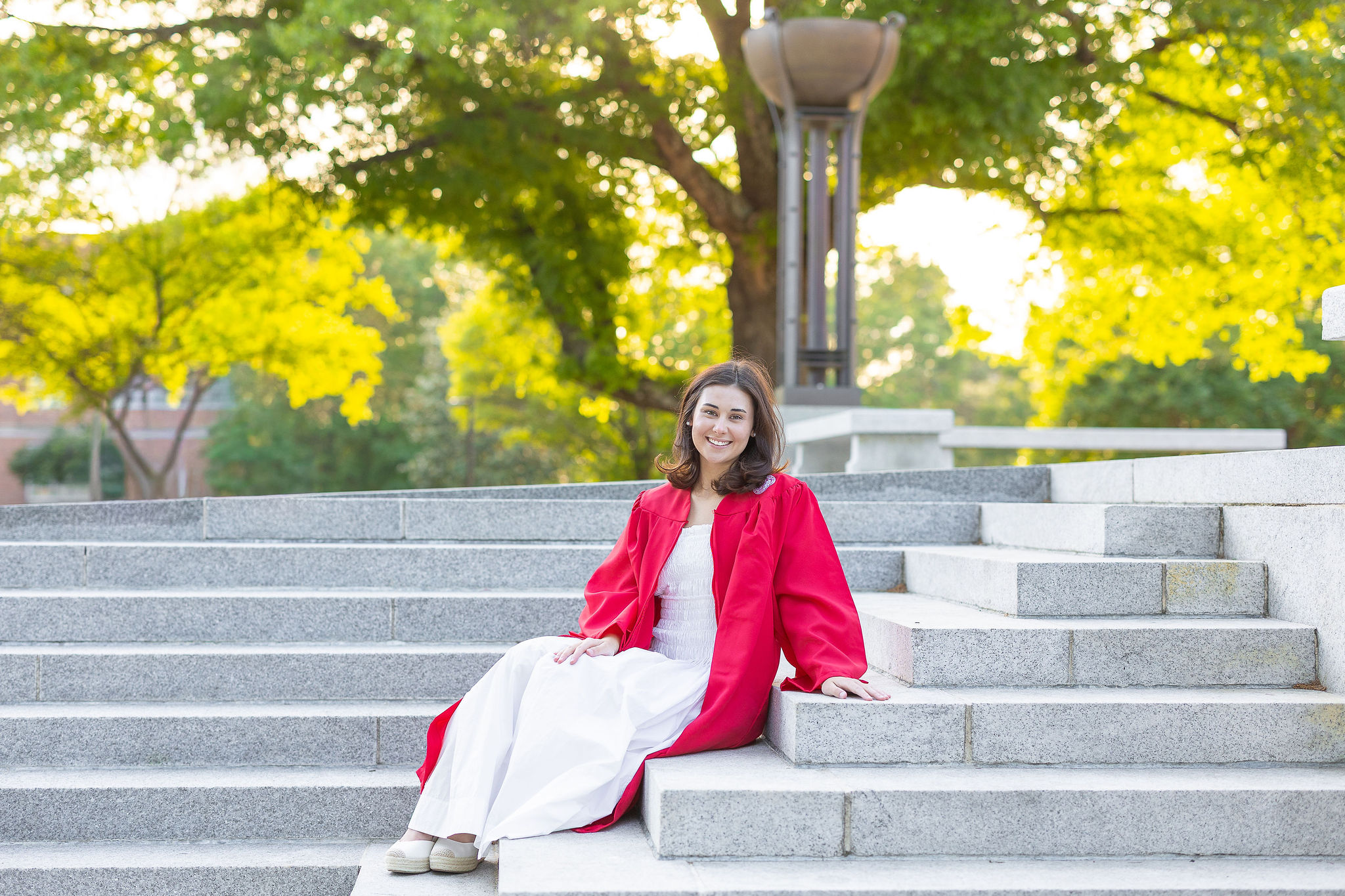 NC State Grad in white long dress and red graduation gown sitting on steps of the NC State belltower